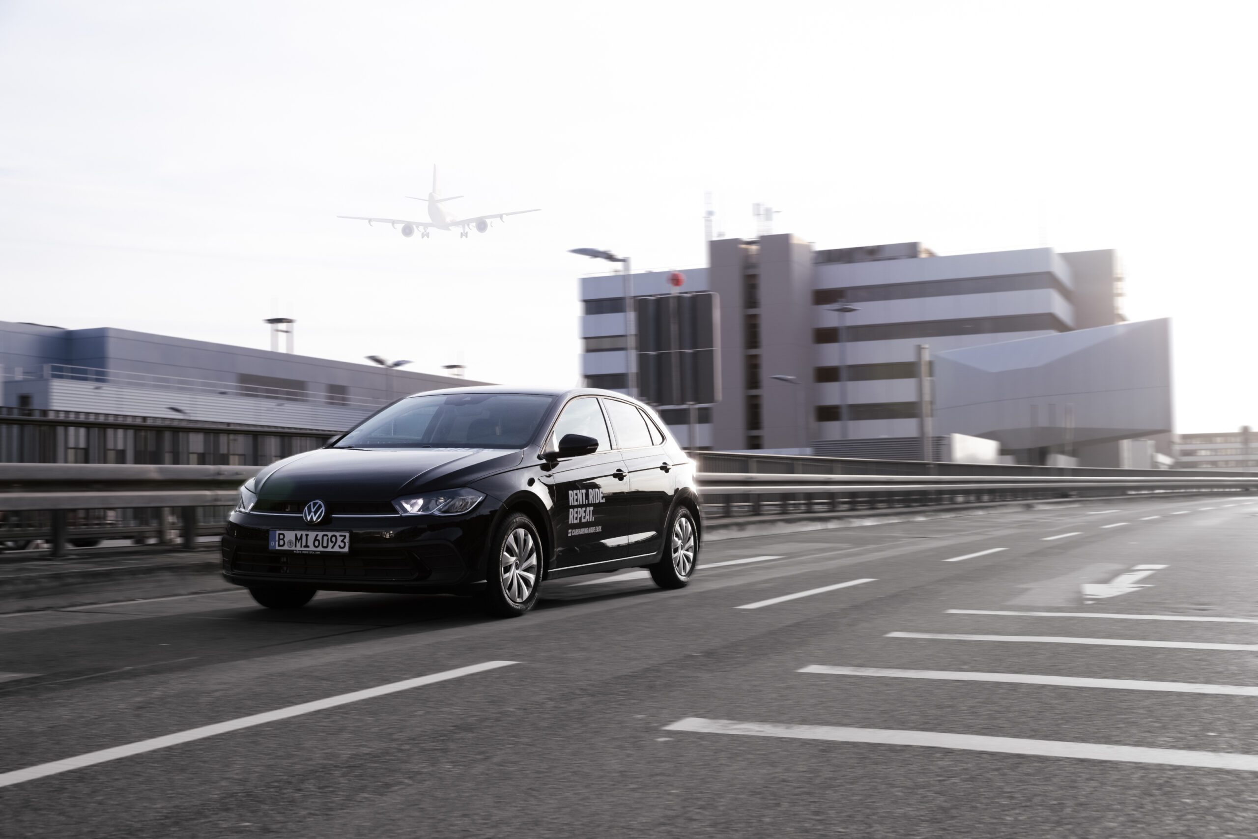 Black Volkswagen Polo with ‘Rent. Ride. Repeat.’ branding driving on a road near Frankfurt Airport with a plane in the background.